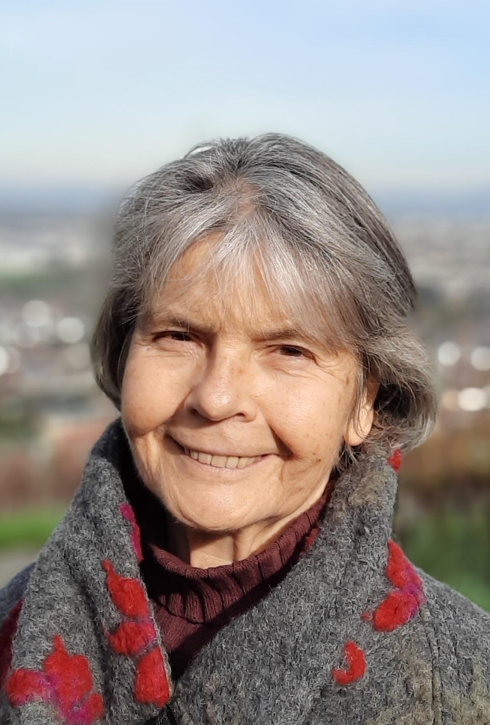 Regina Weiser lächelt im Freien, mit einem grauen Schal mit roten Akzenten.